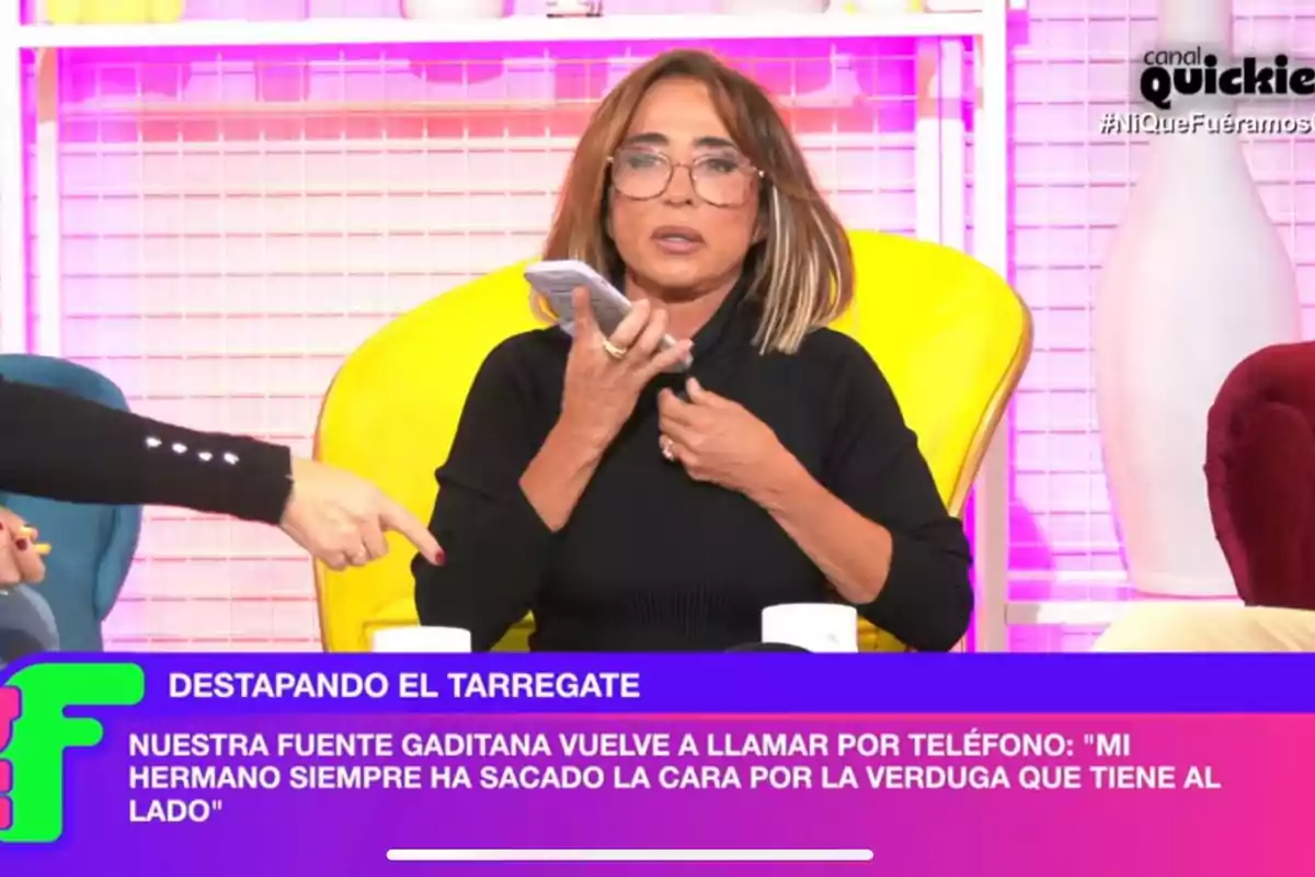 María Patiño con gafas sostiene un teléfono mientras otra persona señala hacia ella en un programa de televisión con un fondo colorido y un texto en la parte inferior que menciona 