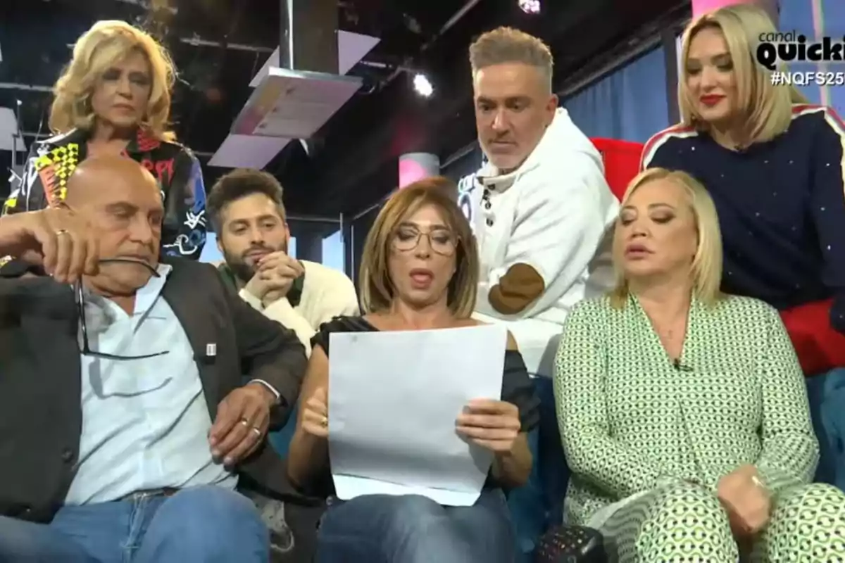 Un grup de persones està reunit en un estudi de televisió, observant atentament un document que sosté una dona al centre.