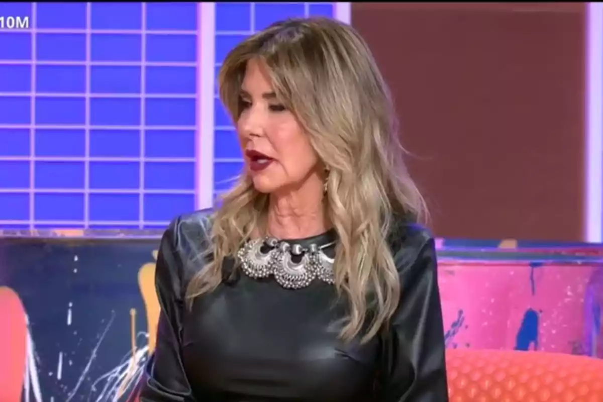 Marisa Martín-Blázquez con cabello rubio y ondulado usando un vestido negro con un collar decorativo en un set de televisión colorido. Marisa Martín-Blázquez con cabello rubio y ondulado usando un vestido negro con un collar decorativo en un set de televisión colorido.
