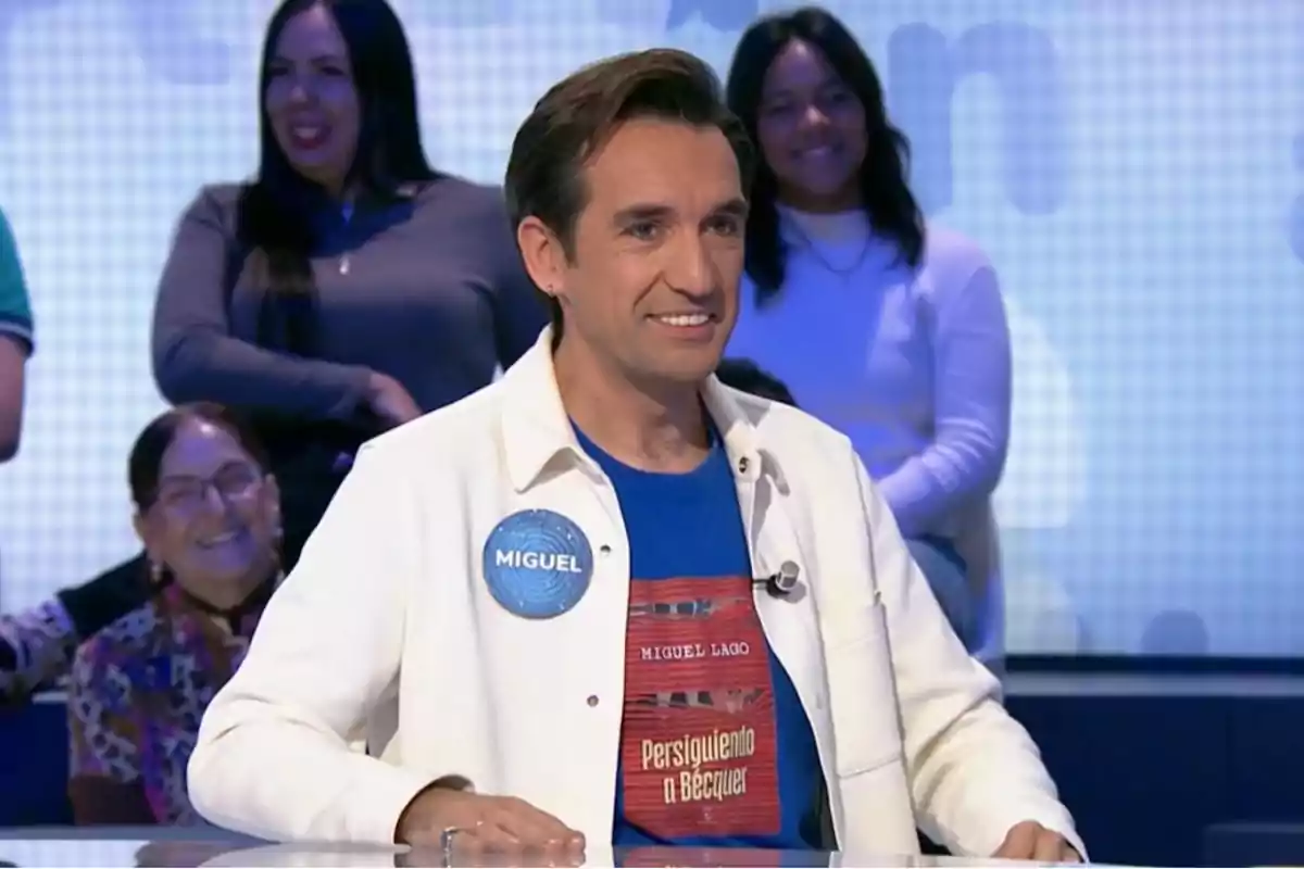 Un home amb una camisa blanca i una samarreta blava somriu mentre està assegut en un programa de televisió amb persones al fons. Un home amb una camisa blanca i una samarreta blava somriu mentre està assegut en un programa de televisió amb persones al fons.