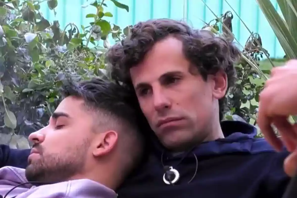Óscar Landa y Miguel Frigenti están sentados juntos, Miguel con la cabeza apoyada en el hombro de Óscar, en un entorno al aire libre con plantas de fondo.