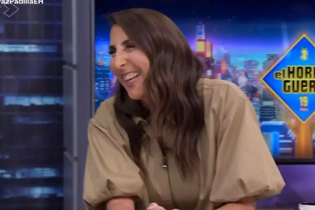 Una persona somrient en un programa de televisió amb un fons acolorit i un logotip visible.