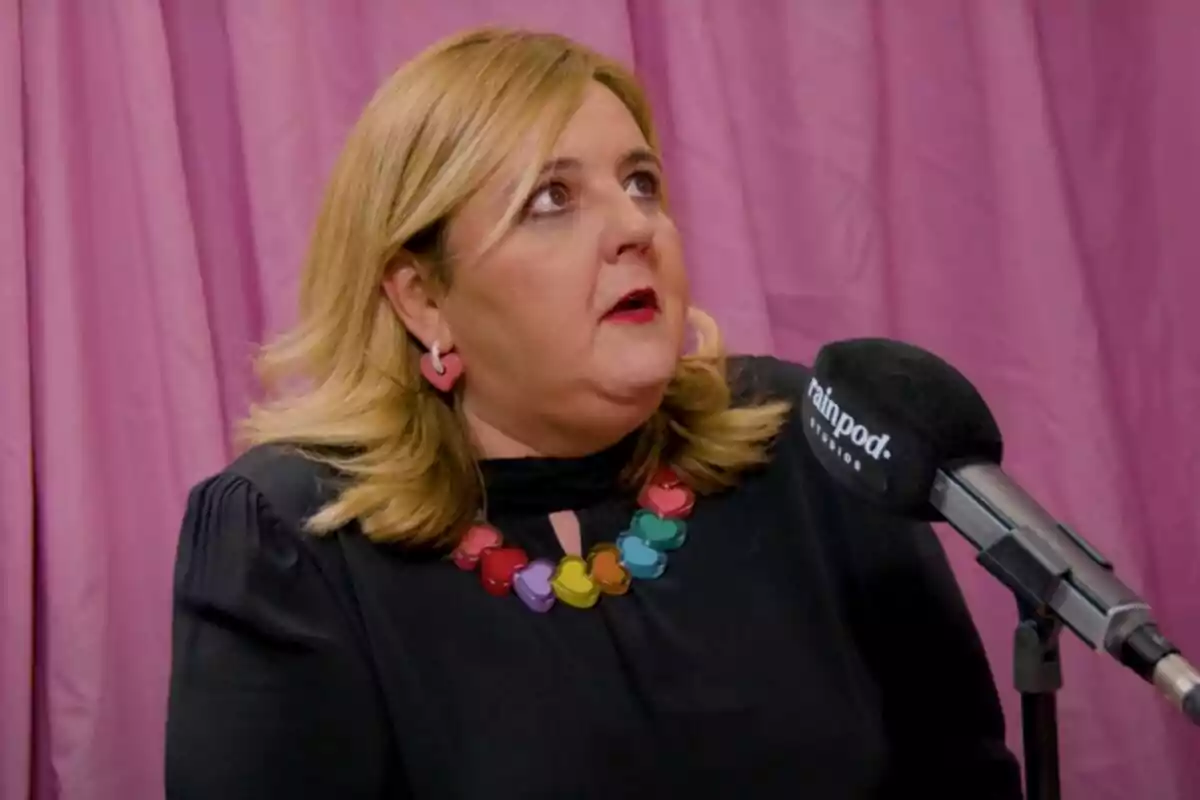 Pilar Vidal con cabello rubio y aretes de corazón rosa está hablando frente a un micrófono con un fondo de cortinas rosas. Lleva un collar de corazones de colores. Pilar Vidal con cabello rubio y aretes de corazón rosa está hablando frente a un micrófono con un fondo de cortinas rosas. Lleva un collar de corazones de colores.