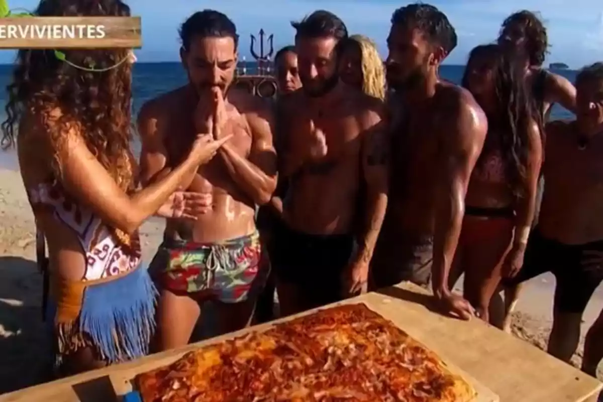 Un grup de persones a la platja observa una gran pizza sobre una taula mentre una dona interactua amb un dels homes. Un grup de persones a la platja observa una gran pizza sobre una taula mentre una dona interactua amb un dels homes.