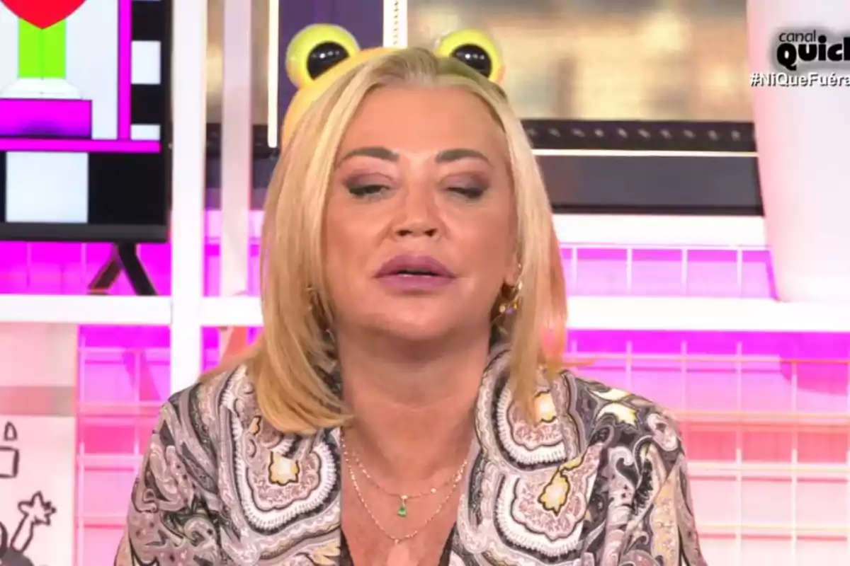 Belén Esteban con cabello rubio y expresión relajada en un set de televisión con luces rosadas y un fondo decorativo.