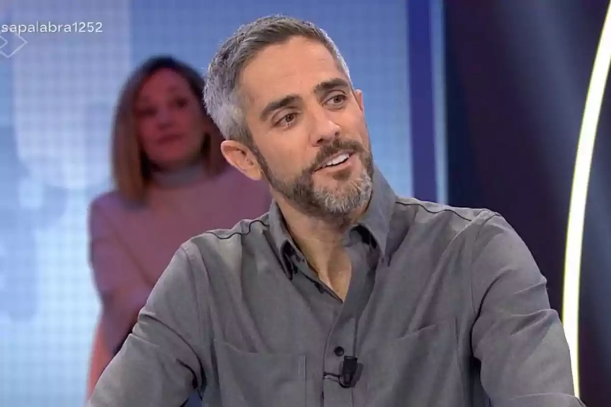 Un home amb barba i camisa grisa somriu mentre està assegut en un estudi de televisió amb una persona desenfocada al fons. Un home amb barba i camisa grisa somriu mentre està assegut en un estudi de televisió amb una persona desenfocada al fons.