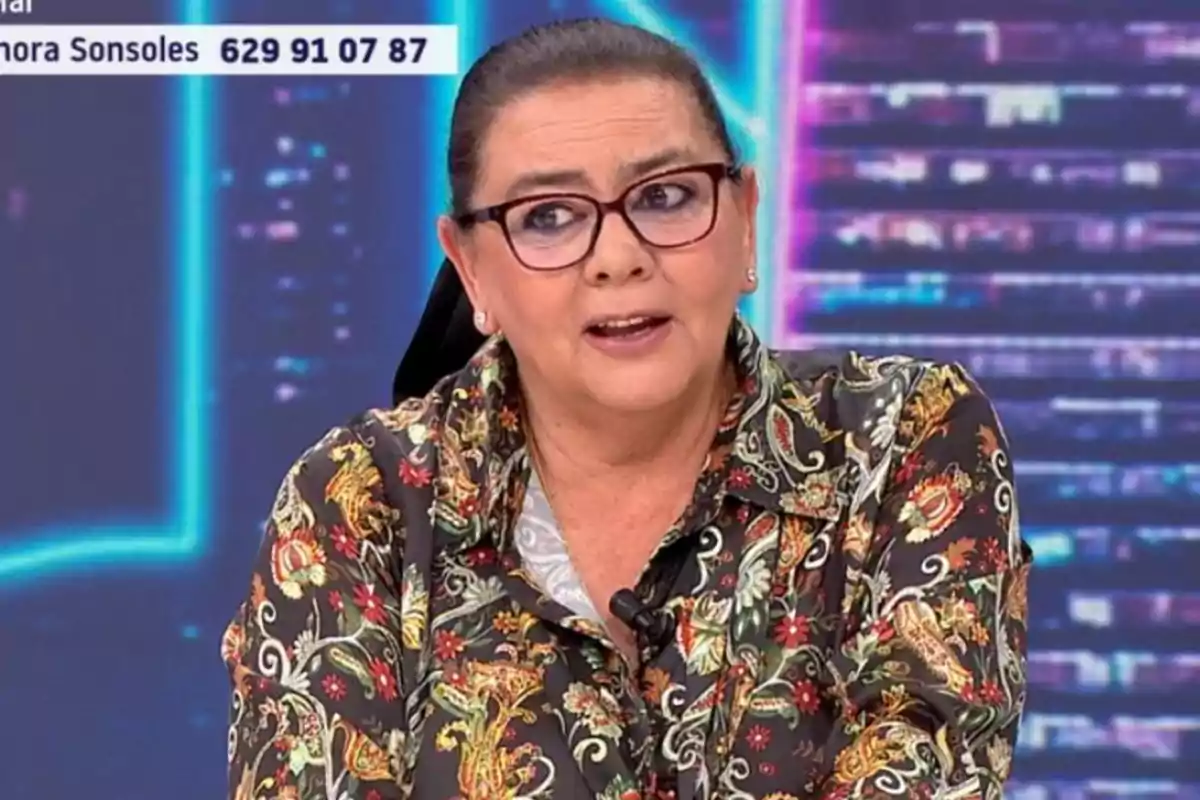 Una persona amb ulleres i camisa d'estampat floral està parlant en un programa de televisió amb un fons de llums i un número de telèfon visible a la cantonada superior esquerra.