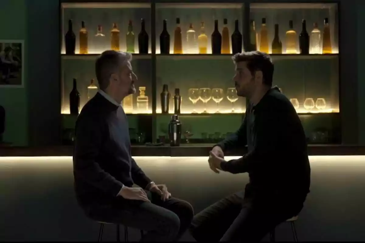 Dos personas sentadas frente a frente en un bar con estantes iluminados llenos de botellas y copas detrás de ellos. Dos personas sentadas frente a frente en un bar con estantes iluminados llenos de botellas y copas detrás de ellos.