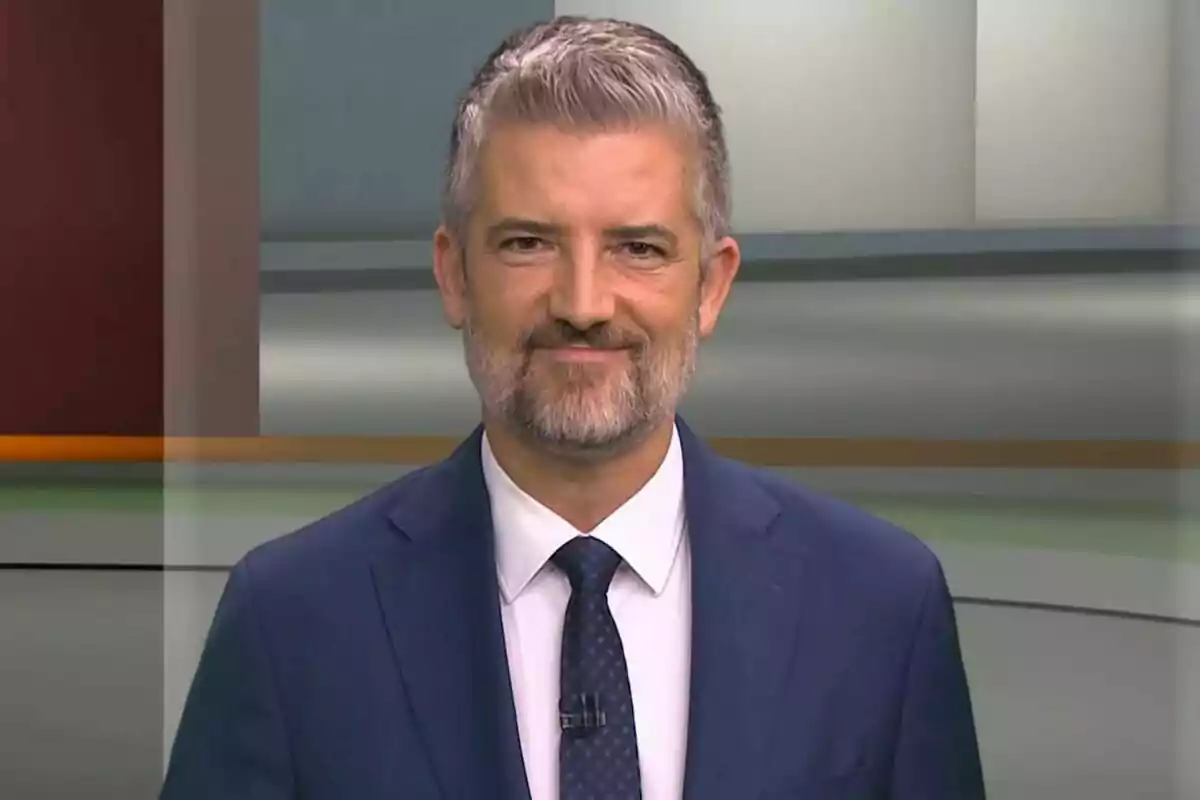Un home amb barba i cabell canós porta un vestit blau i corbata, somrient davant d'un fons d'estudi.