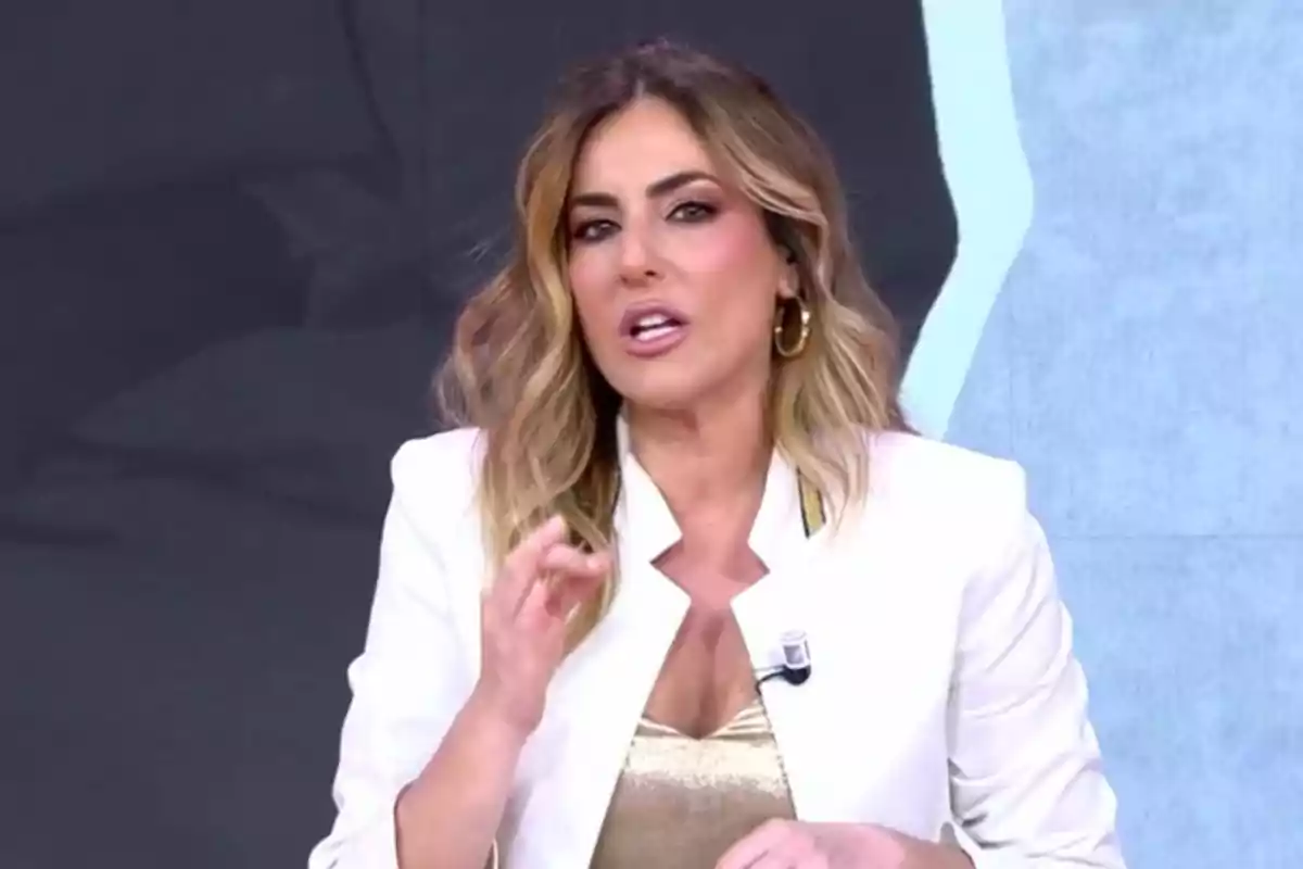 Verónica Dulanto con cabello rubio y ondulado, vistiendo un blazer blanco y blusa dorada, hablando frente a un micrófono.