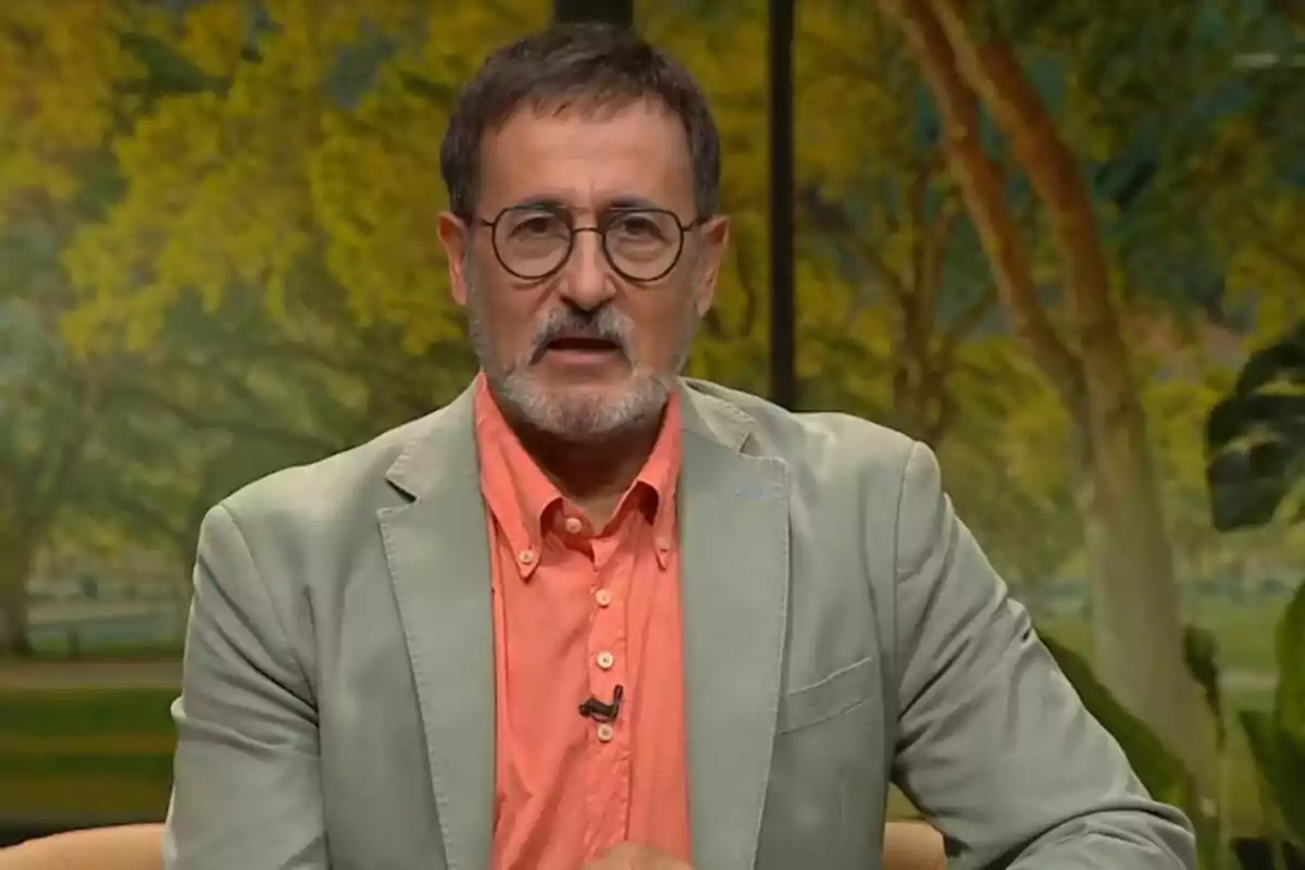 Hombre con gafas y barba, vestido con una chaqueta verde claro y camisa naranja, sentado frente a un fondo de árboles. Hombre con gafas y barba, vestido con una chaqueta verde claro y camisa naranja, sentado frente a un fondo de árboles.