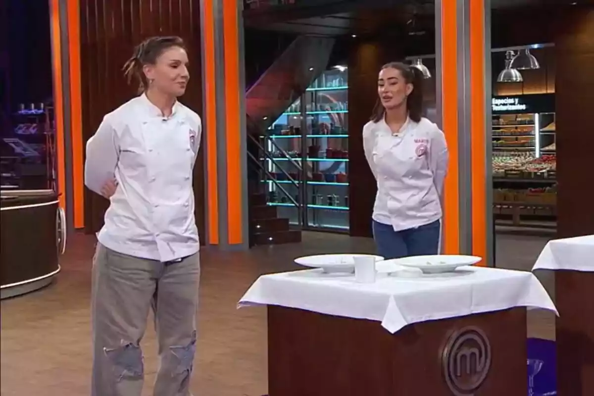 Inés Hernand y Marina Rivers en MasterChef Celebrity están de pie frente a una mesa con platos vacíos, vistiendo chaquetas de chef blancas. Inés Hernand y Marina Rivers en MasterChef Celebrity están de pie frente a una mesa con platos vacíos, vistiendo chaquetas de chef blancas.