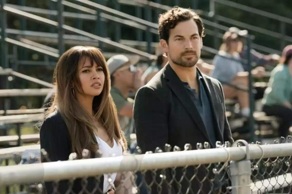 Fotografía de Vanessa Morgan y Giacomo Gianniotti de pie frente a una cerca en un estadio al aire libre en la serie Infiltrada Fotografía de Vanessa Morgan y Giacomo Gianniotti de pie frente a una cerca en un estadio al aire libre en la serie Infiltrada