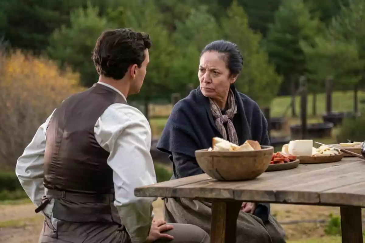 Xavi Lock y Inma Perez-Quirós sentados en una mesa de madera al aire libre con platos de comida, rodeados de un paisaje natural con árboles en La Promesa. Xavi Lock y Inma Perez-Quirós sentados en una mesa de madera al aire libre con platos de comida, rodeados de un paisaje natural con árboles en La Promesa.