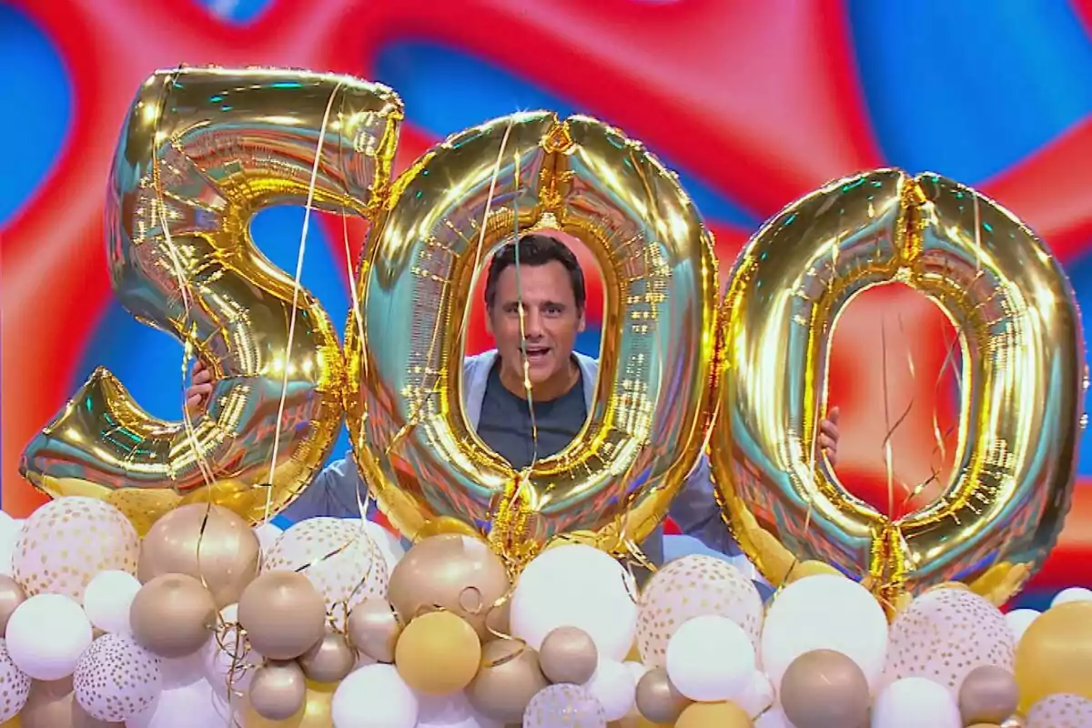 Ion Aramendi sosteniendo globos dorados con el número 500 rodeado de globos blancos y dorados sobre un fondo colorido en Reacción en Cadena. Ion Aramendi sosteniendo globos dorados con el número 500 rodeado de globos blancos y dorados sobre un fondo colorido en Reacción en Cadena.