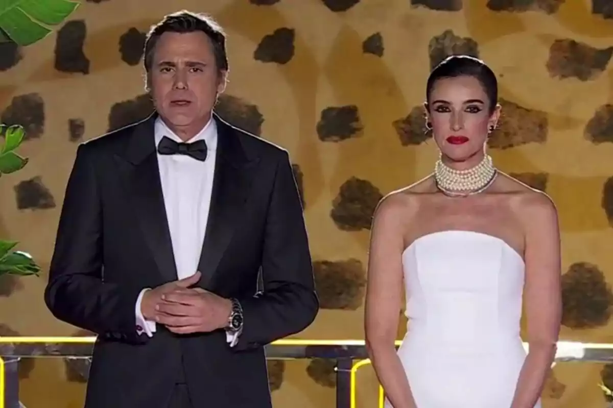 Ion Aramendi y Blanca Romero elegantemente vestidas de pie frente a un fondo decorativo con patrones oscuros y hojas verdes en las Campanadas.