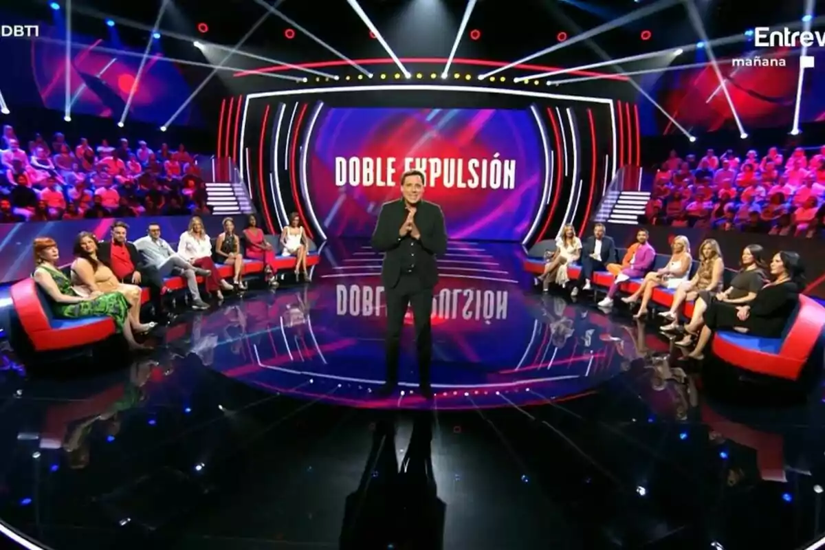 Ion Aramendi amb vestit fosc està parlant al plató de Gran Hermano al costat dels col·laboradors amb una pantalla que diu Ion Aramendi amb vestit fosc està parlant al plató de Gran Hermano al costat dels col·laboradors amb una pantalla que diu