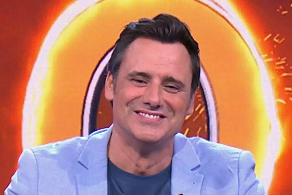 Ion Aramendi sonriente con chaqueta azul claro frente a un fondo naranja brillante en Reacción en Cadena. Ion Aramendi sonriente con chaqueta azul claro frente a un fondo naranja brillante en Reacción en Cadena.
