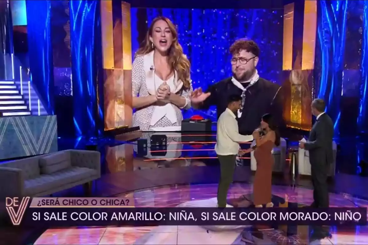 De Viernes con un escenario iluminado en tonos azules y dorados, donde Asraf Beno e Isa Pantoja está de pie frente a Santi Acosta, mientras en una pantalla grande se ven Beatriz Archidona y un amigo, y en la parte inferior hay un texto sobre la revelación de género de un bebé. De Viernes con un escenario iluminado en tonos azules y dorados, donde Asraf Beno e Isa Pantoja está de pie frente a Santi Acosta, mientras en una pantalla grande se ven Beatriz Archidona y un amigo, y en la parte inferior hay un texto sobre la revelación de género de un bebé.