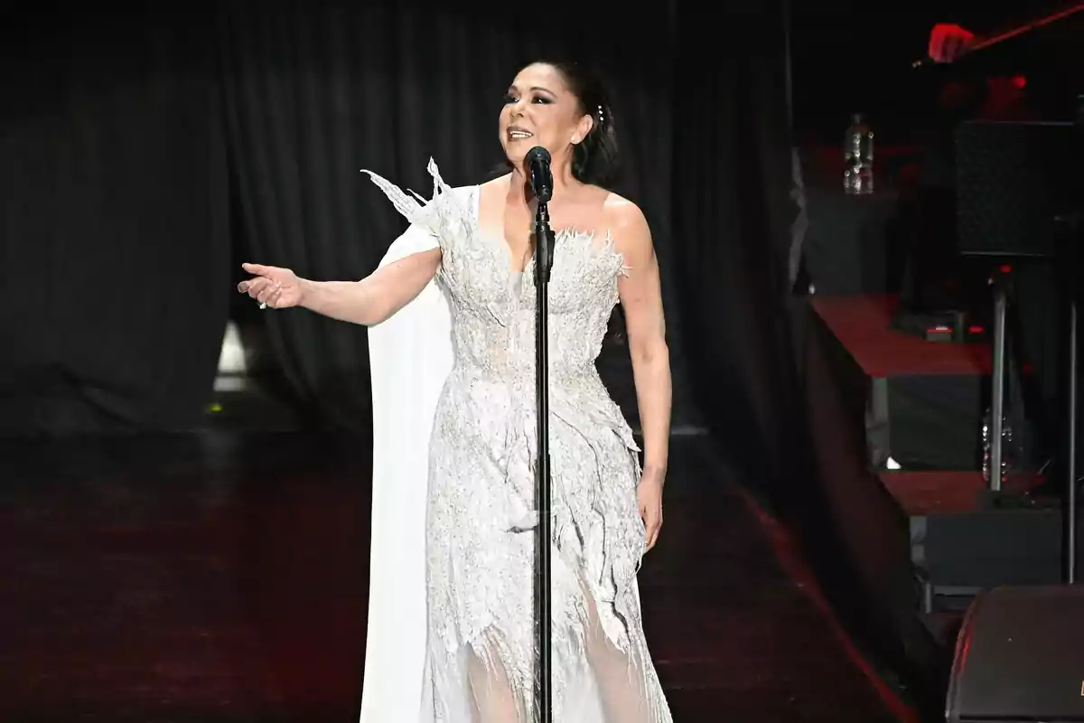 Isabel Pantoja con un vestido blanco elegante canta en un escenario iluminado. Isabel Pantoja con un vestido blanco elegante canta en un escenario iluminado.