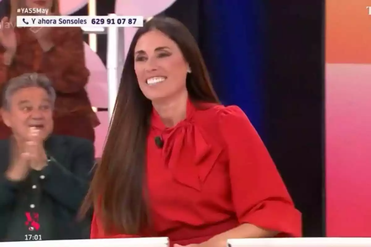 Isabel Rábago somrient amb un vestit vermell està asseguda a 'Y ahora Sonsoles', mentre una persona aplaudeix al fons.