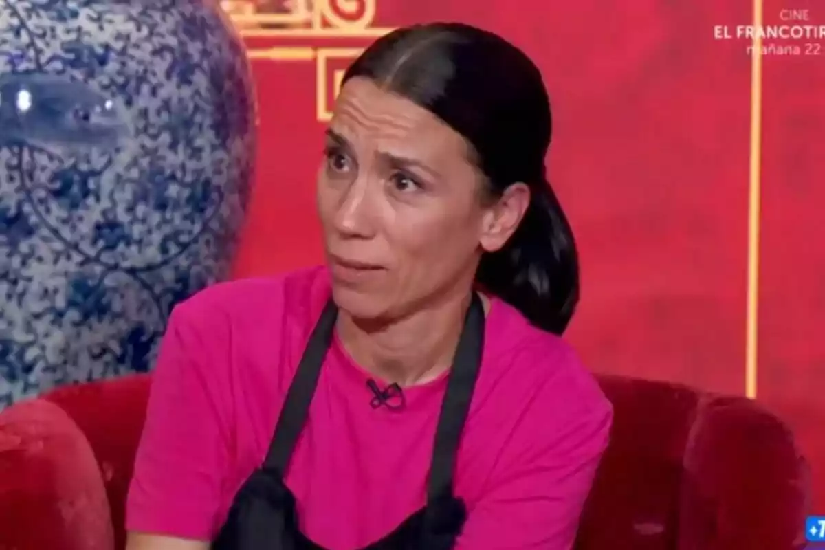 Itziar Miranda con expresión de sorpresa o confusión, vestida con camiseta rosa y delantal negro, sentada en un sofá rojo con un jarrón azul decorativo al fondo en MasterChef Celebrity. Itziar Miranda con expresión de sorpresa o confusión, vestida con camiseta rosa y delantal negro, sentada en un sofá rojo con un jarrón azul decorativo al fondo en MasterChef Celebrity.