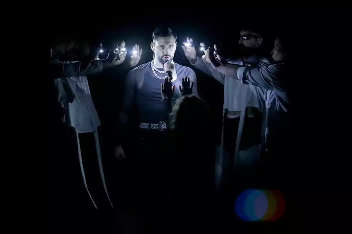 JKbello canta en un escenario oscuro rodeado de personas que sostienen luces brillantes en el Benidorm Fest. JKbello canta en un escenario oscuro rodeado de personas que sostienen luces brillantes en el Benidorm Fest.