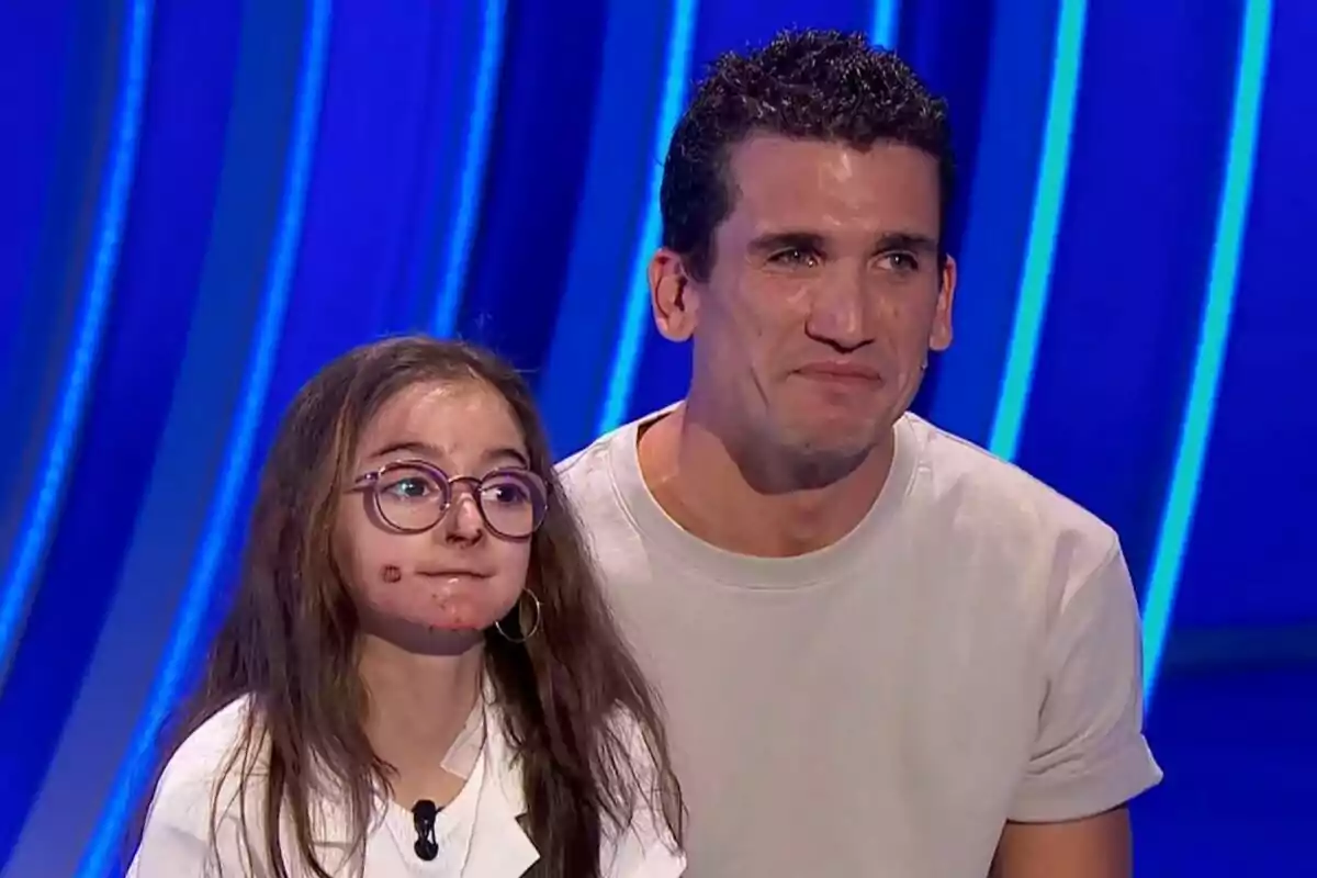Una niña con gafas y Jaime Lorente sonríen en un escenario con fondo azul en Hay una cosa que te quiero decir. Una niña con gafas y Jaime Lorente sonríen en un escenario con fondo azul en Hay una cosa que te quiero decir.