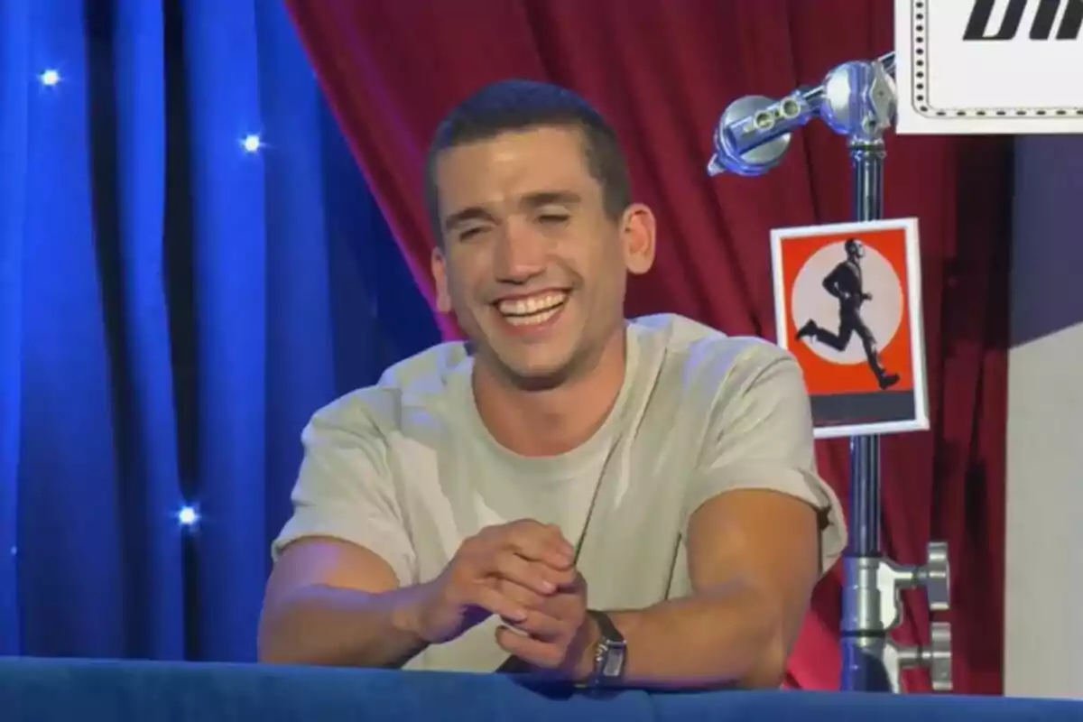 Jaime Lorente sonriente está sentado frente a un fondo con cortinas azules y rojas y una señal de advertencia con una figura corriendo en El Gran Show. Jaime Lorente sonriente está sentado frente a un fondo con cortinas azules y rojas y una señal de advertencia con una figura corriendo en El Gran Show.