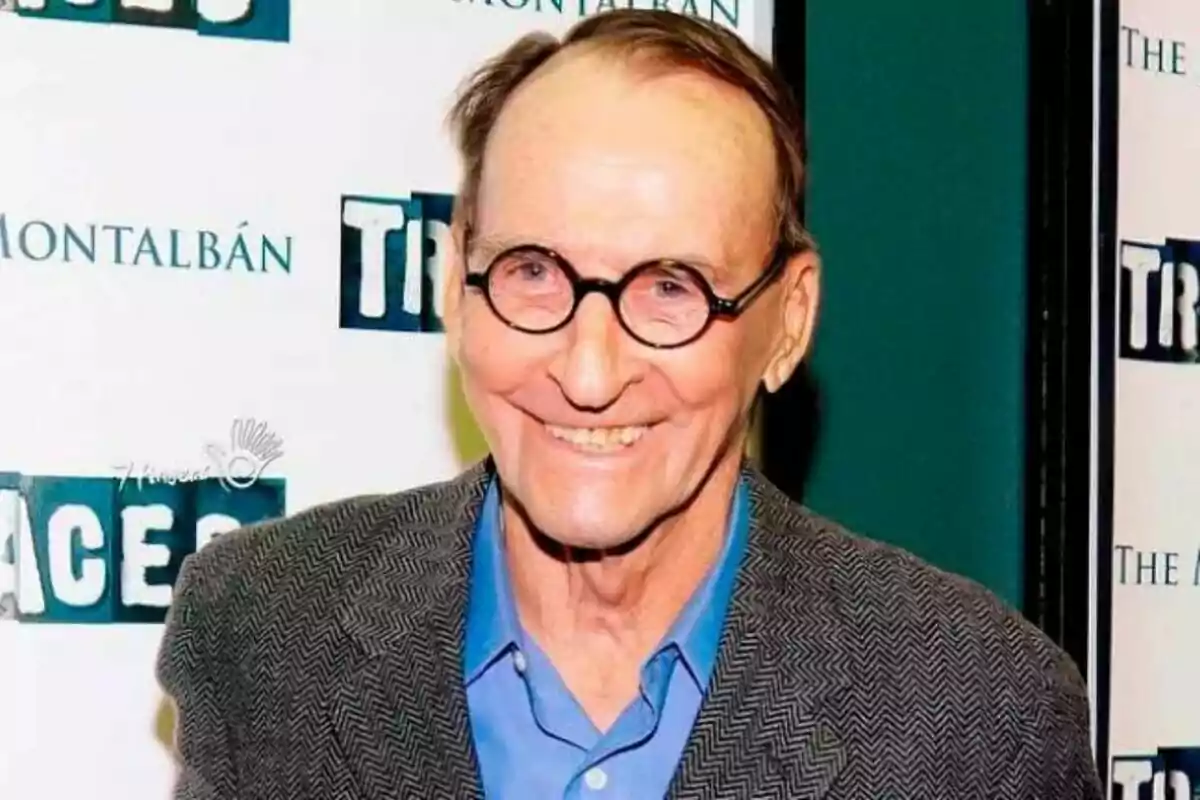Fotografía de James B. Sikking con gafas redondas y sonrisa, vestido con camisa azul y chaqueta gris, posando frente a un fondo con logotipos. Fotografía de James B. Sikking con gafas redondas y sonrisa, vestido con camisa azul y chaqueta gris, posando frente a un fondo con logotipos.