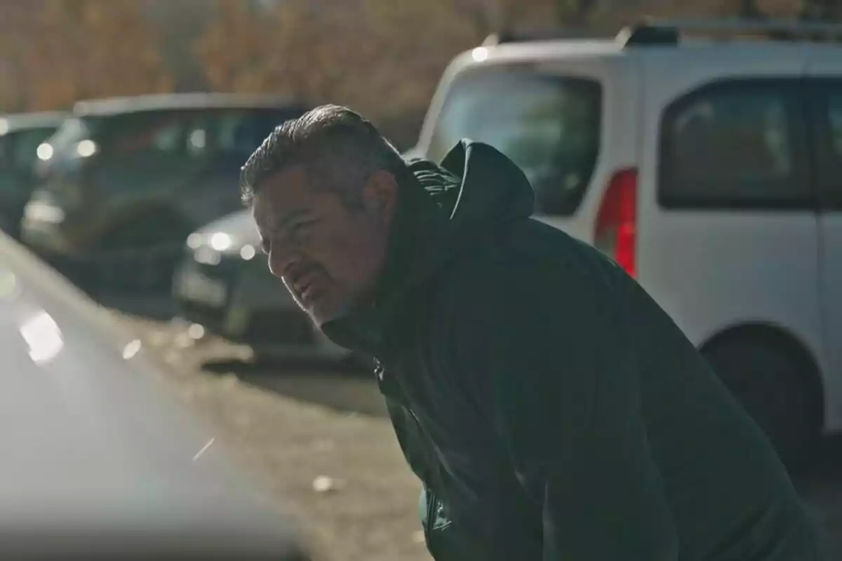 Jalis de la Serna con chaqueta oscura está inclinado cerca de un automóvil en un estacionamiento, con varios vehículos en el fondo en Apatrullando.