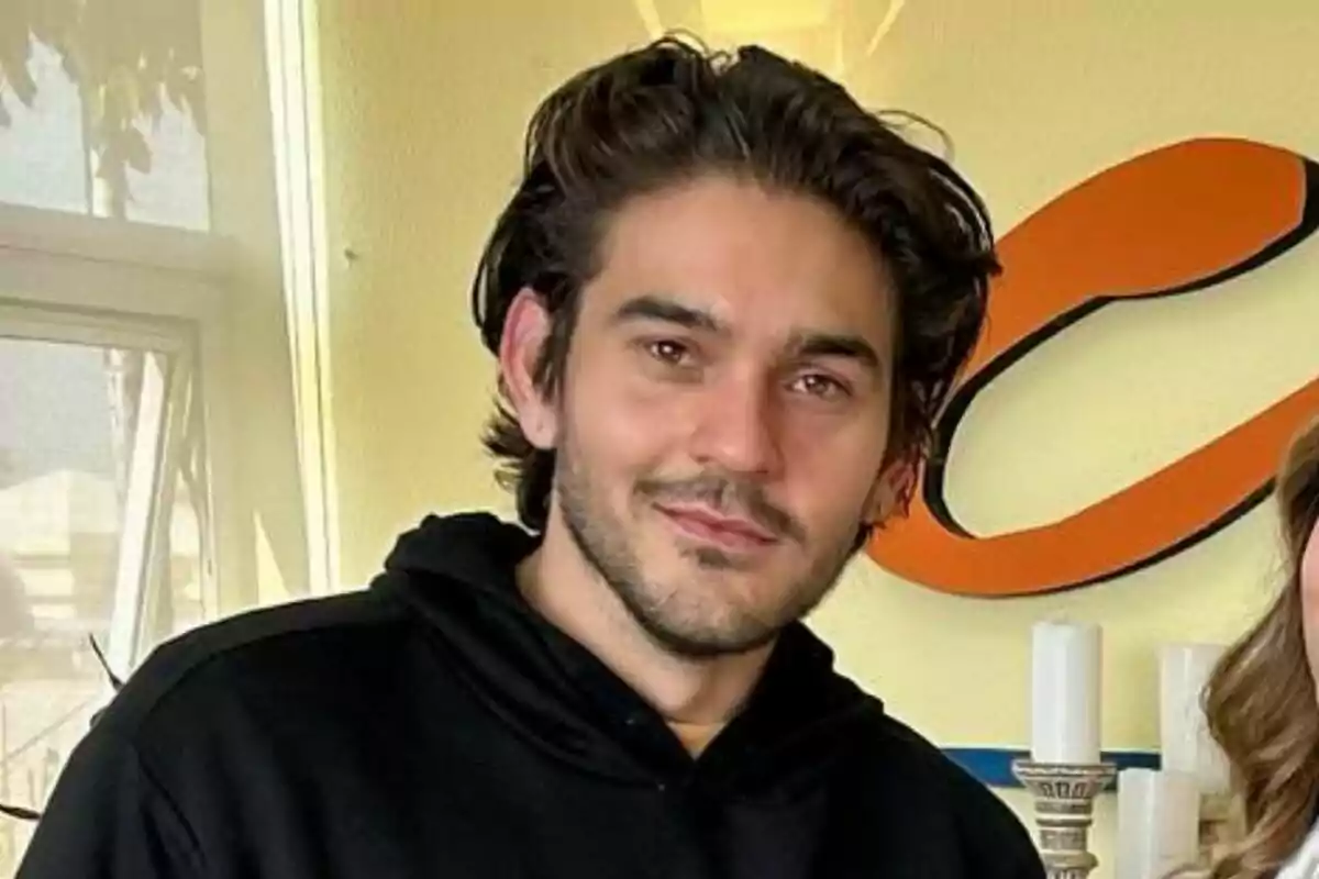 James Hollcroft con cabello oscuro y barba ligera, vistiendo una sudadera negra, posando frente a una pared amarilla con una ventana y una decoración en forma de letra James Hollcroft con cabello oscuro y barba ligera, vistiendo una sudadera negra, posando frente a una pared amarilla con una ventana y una decoración en forma de letra
