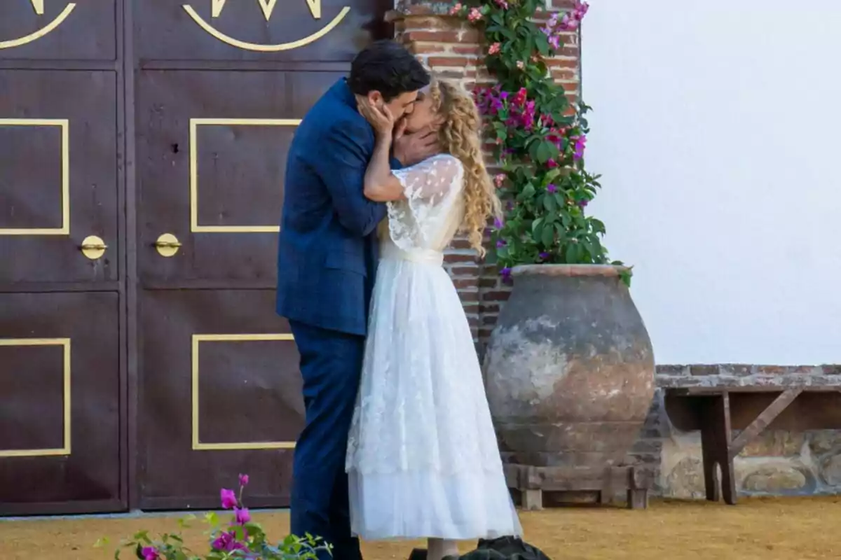Jana y Manuel se besa apasionadamente frente a una puerta de madera con detalles dorados, junto a una maceta grande y flores en La Promesa. Jana y Manuel se besa apasionadamente frente a una puerta de madera con detalles dorados, junto a una maceta grande y flores en La Promesa.