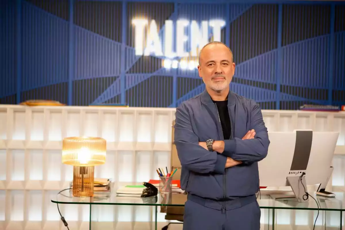 Javier Gutiérrez dret amb els braços creuats davant d'un escriptori amb una làmpada i un ordinador en un entorn modern amb un rètol que diu 
