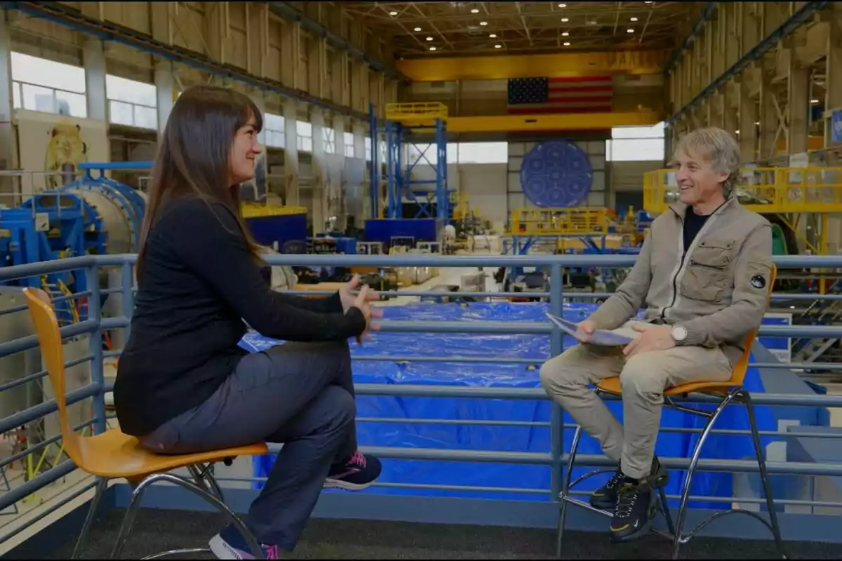 Jesús Calleja y otra mujer sentadas en sillas conversan en el interior de una gran instalación industrial con maquinaria y una bandera estadounidense al fondo en Calleja en el espacio. Jesús Calleja y otra mujer sentadas en sillas conversan en el interior de una gran instalación industrial con maquinaria y una bandera estadounidense al fondo en Calleja en el espacio.