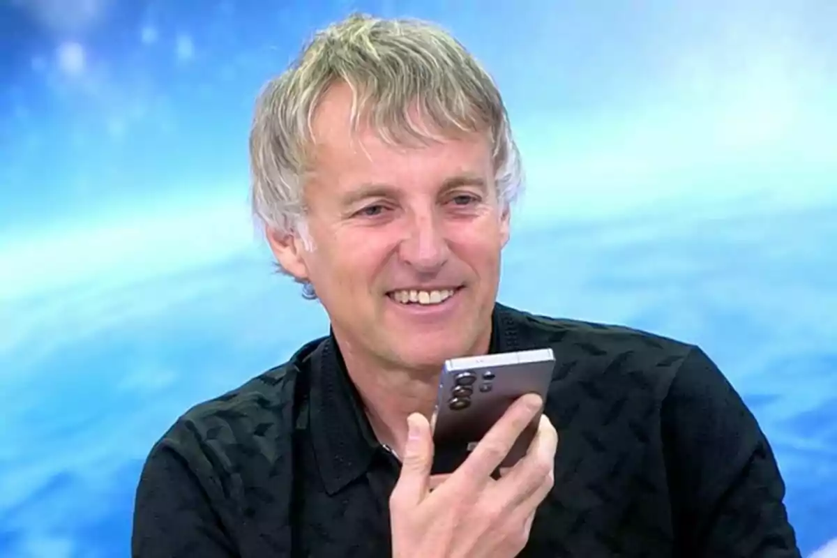 Jesús Calleja somrient mentre sosté un telèfon mòbil en un entorn amb fons blau a 'El programa de Ana Rosa'.