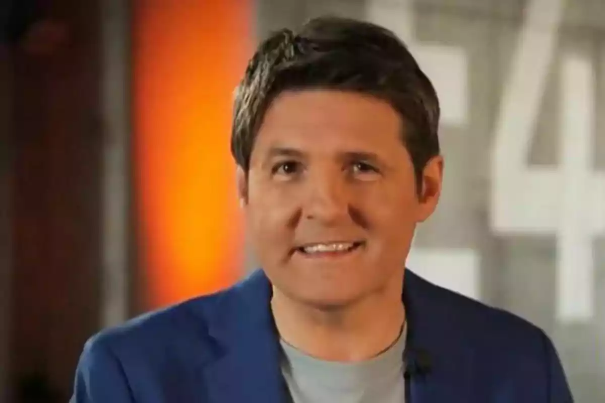 Jesús Cintora con chaqueta azul y camiseta gris sonriendo frente a un fondo difuminado en tonos naranjas y grises en la promo de Malas Lenguas.