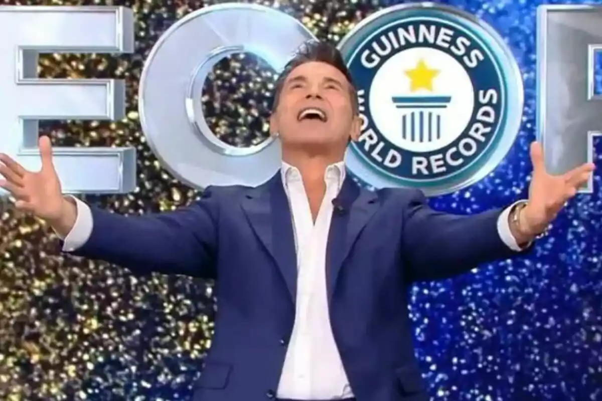 Jesús Vázquez amb vestit blau i camisa blanca aixeca els braços i somriu davant d'un fons amb el logotip de Guinness World Records a La Nit dels Rècords.