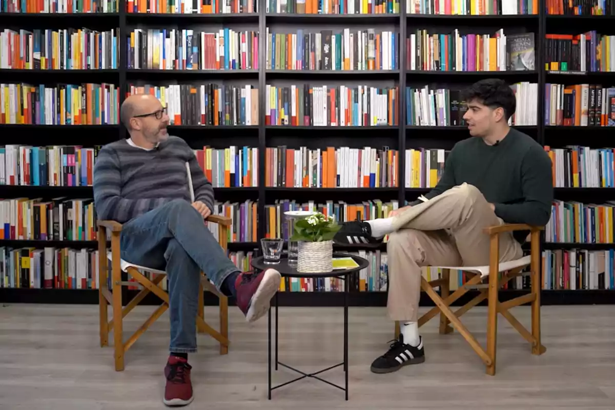 Jon Garcia i Joan María Pou asseguts en cadires de fusta conversant davant d'una taula petita amb una planta, en un entorn envoltat d'estanteries plenes de llibres.