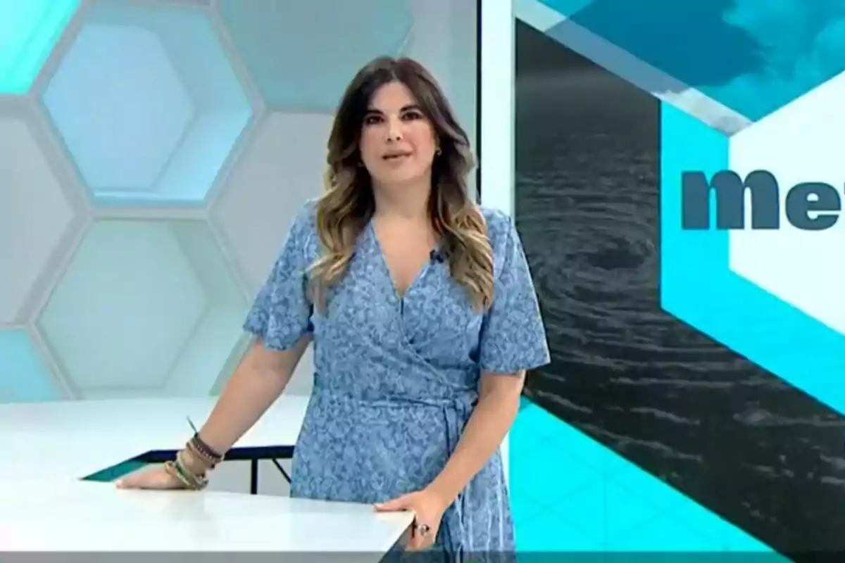 Joanna Ivars de pie en un estudio de televisión con un vestido azul y fondo geométrico.