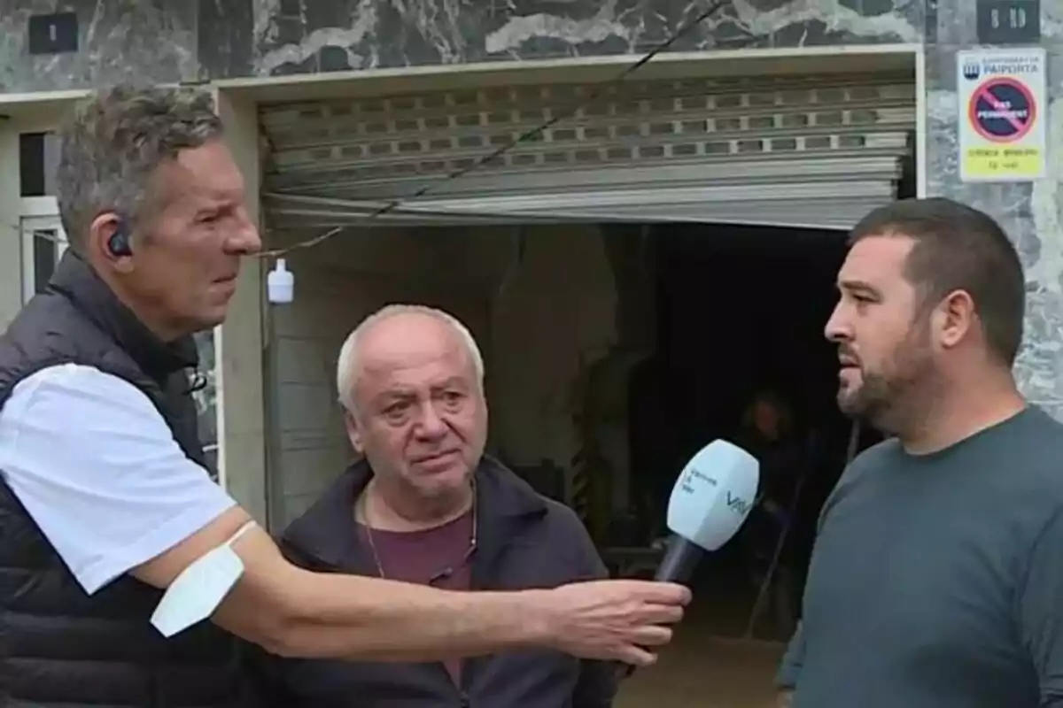Joaquín Prat sostiene un micrófono de Vamos a ver mientras entrevista a dos hombres frente a un edificio con una puerta enrollable. Joaquín Prat sostiene un micrófono de Vamos a ver mientras entrevista a dos hombres frente a un edificio con una puerta enrollable.