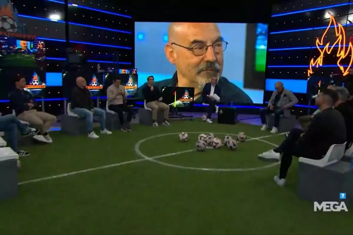 Un grup de persones com Josep Pedrerol està assegut a El Chiringuito de Jugones amb temàtica de futbol, amb un gran retrat de Joaquín Ramos Marcos calb amb ulleres a la pantalla de fons i diverses pilotes de futbol al centre del set.