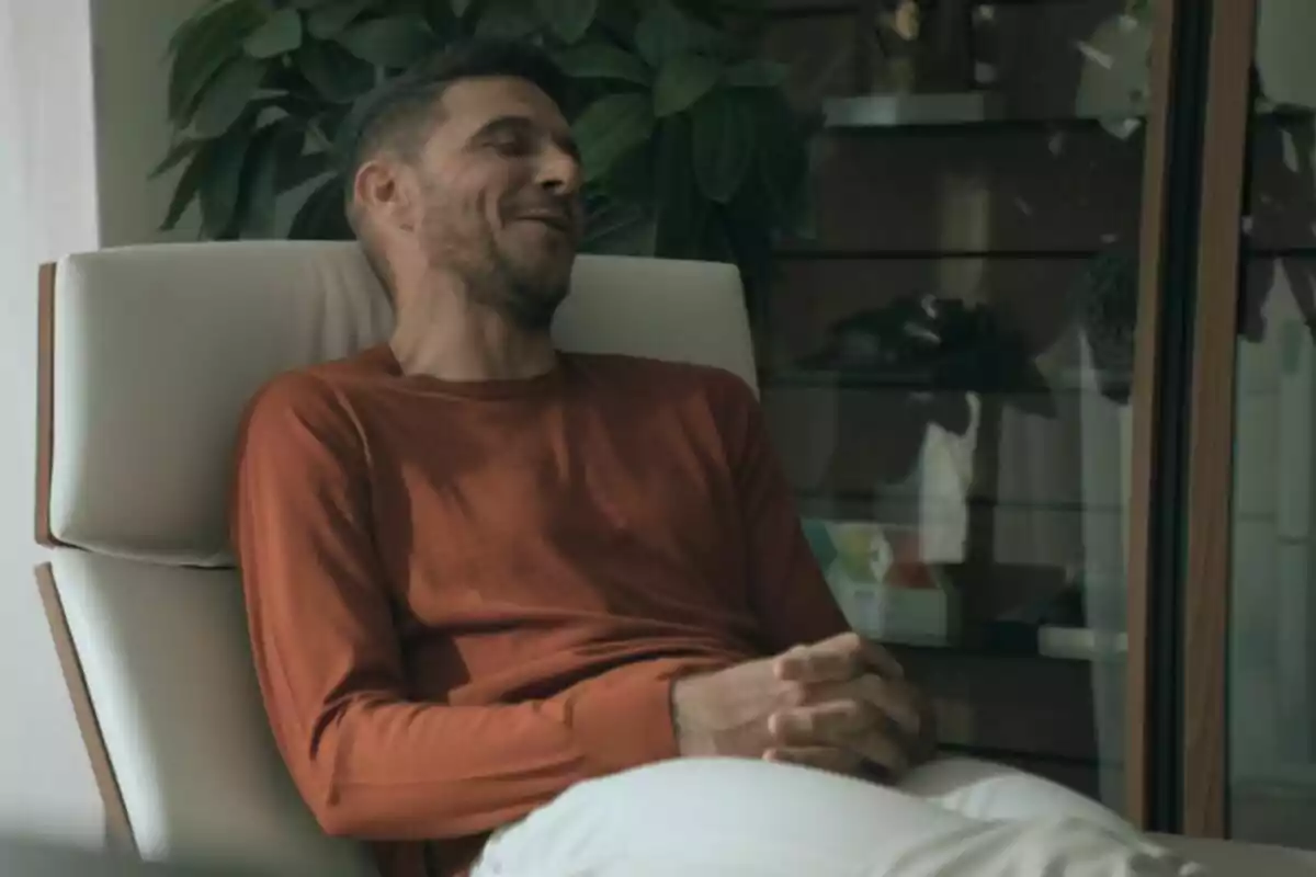 Joaquín Sánchez con barba y cabello corto está sentado en una silla blanca, vistiendo un suéter naranja y pantalones claros, con las manos entrelazadas y una expresión relajada, mientras una planta y un mueble con objetos decorativos se ven al fondo en El Capitán en América. Joaquín Sánchez con barba y cabello corto está sentado en una silla blanca, vistiendo un suéter naranja y pantalones claros, con las manos entrelazadas y una expresión relajada, mientras una planta y un mueble con objetos decorativos se ven al fondo en El Capitán en América.