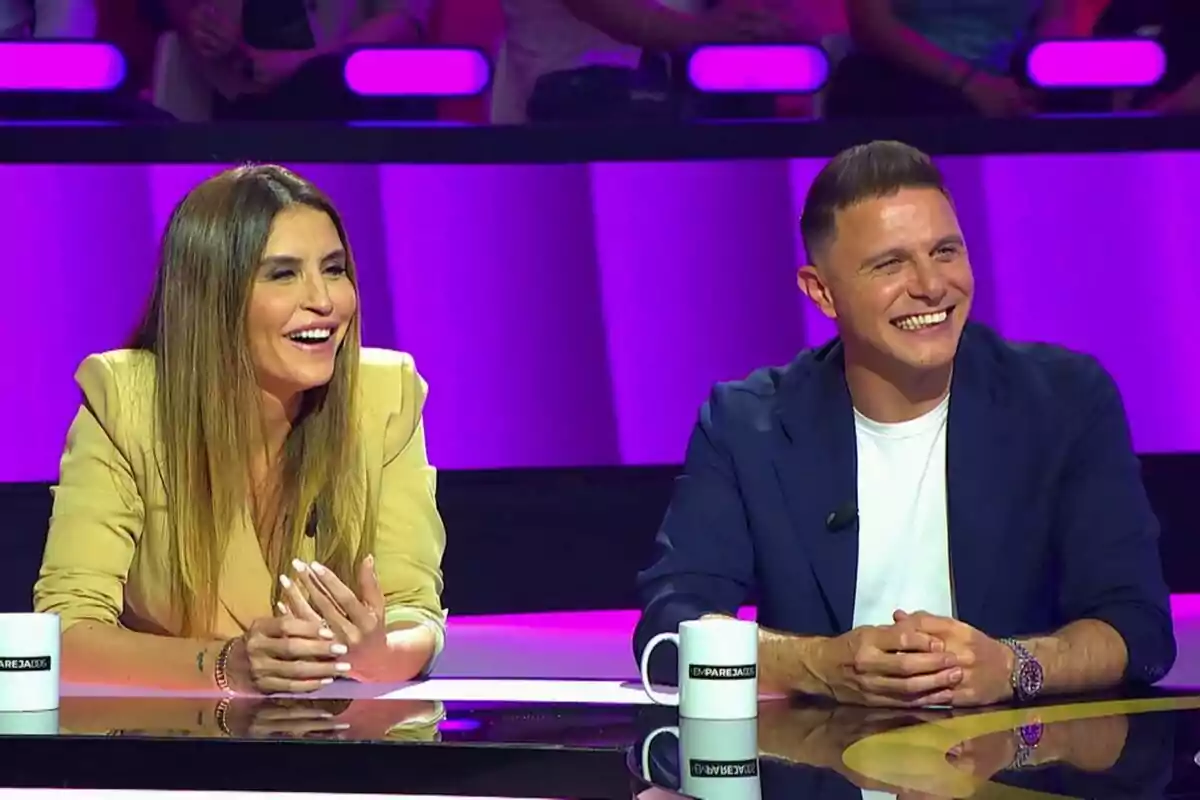 Joaquín Sánchez y Susana Saborido sonrientes sentadas en una mesa con tazas blancas, con un fondo iluminado en tonos morados en Emparejados Joaquín Sánchez y Susana Saborido sonrientes sentadas en una mesa con tazas blancas, con un fondo iluminado en tonos morados en Emparejados