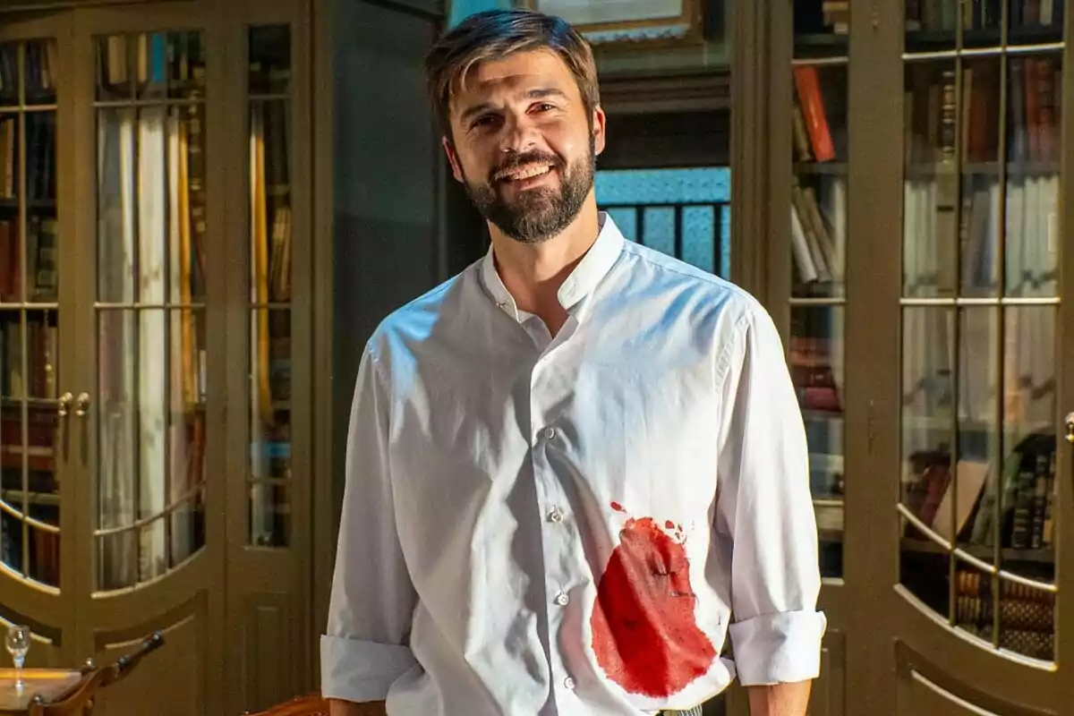 Jordi Coll con barba y camisa blanca manchada de rojo está de pie en una habitación con estanterías llenas de libros en La Promesa. Jordi Coll con barba y camisa blanca manchada de rojo está de pie en una habitación con estanterías llenas de libros en La Promesa.