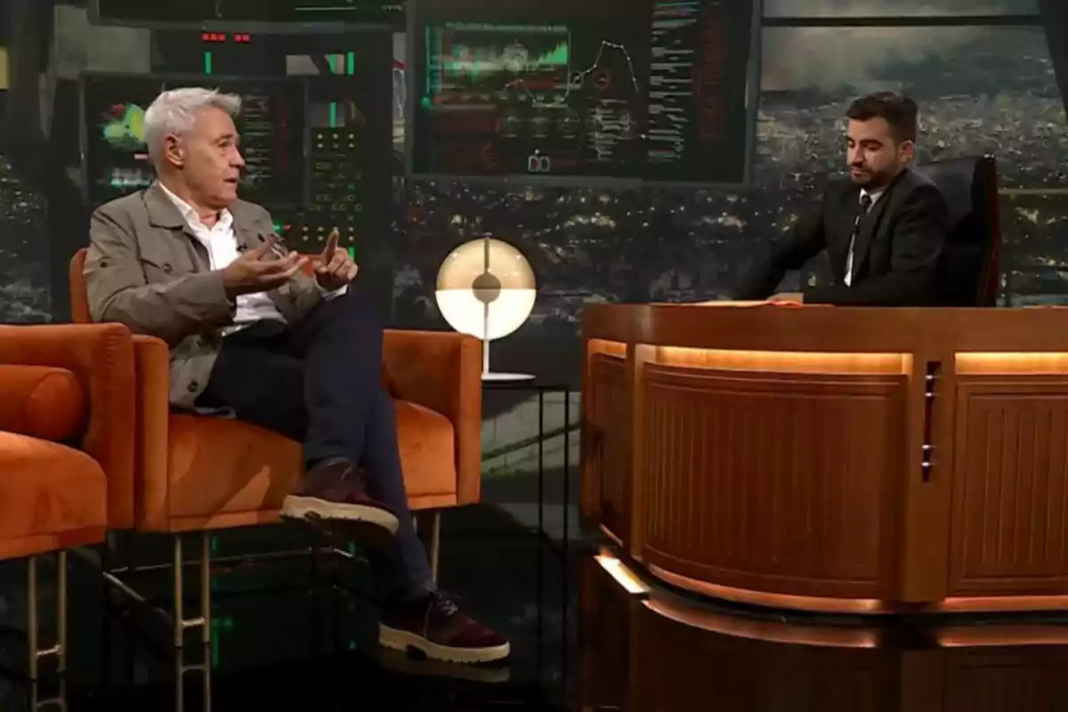 Jordi González i Ricard Ustrell conversen en un plató de televisió de Col·lapse, un assegut en un sofà taronja i l’altre darrere d’un escriptori de fusta amb pantalles i una làmpada de fons.