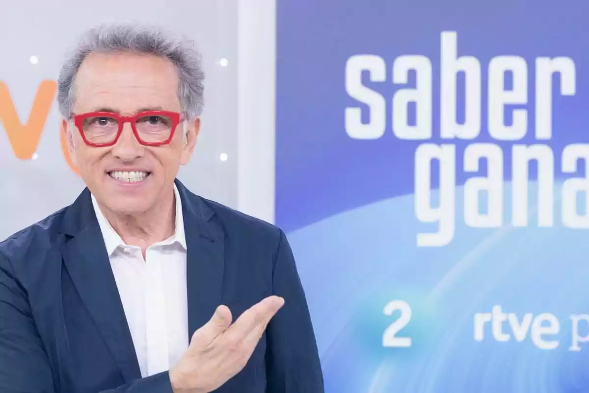 Jordi Hurtado de cabello canoso y gafas rojas sonríe y señala con la mano en un set de televisión con el logo de un programa llamado Saber y Ganar al fondo