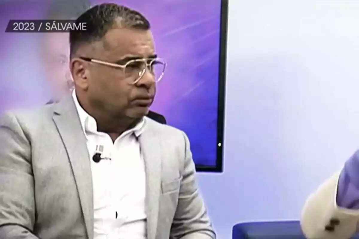 Jorge Javier Vázquez amb ulleres i vestit clar està assegut en un estudi de televisió amb Kiko Hernández difuminat un fons morat i una pantalla darrere en una imatge emesa a De Viernes.