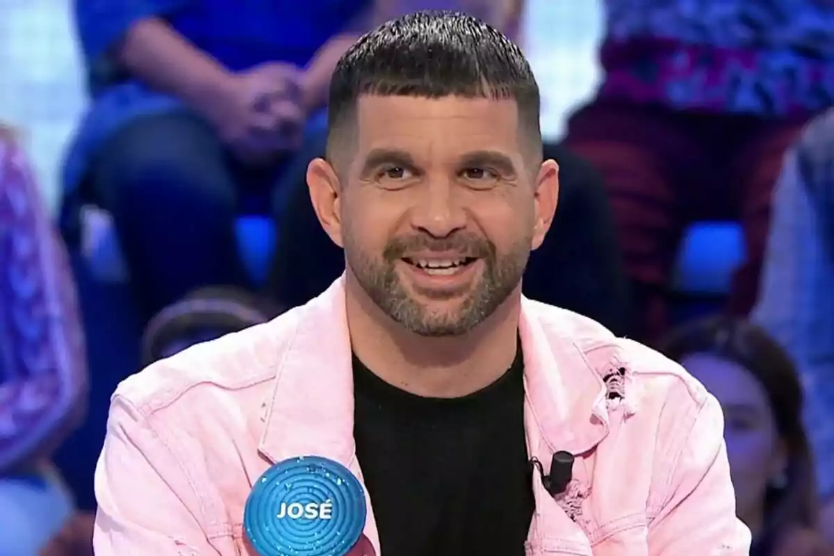 José Álvarez con chaqueta rosa y camiseta negra sonríe mientras está sentado en un programa de televisión, con un micrófono en la solapa y un público desenfocado al fondo en Pasapalabra. José Álvarez con chaqueta rosa y camiseta negra sonríe mientras está sentado en un programa de televisión, con un micrófono en la solapa y un público desenfocado al fondo en Pasapalabra.