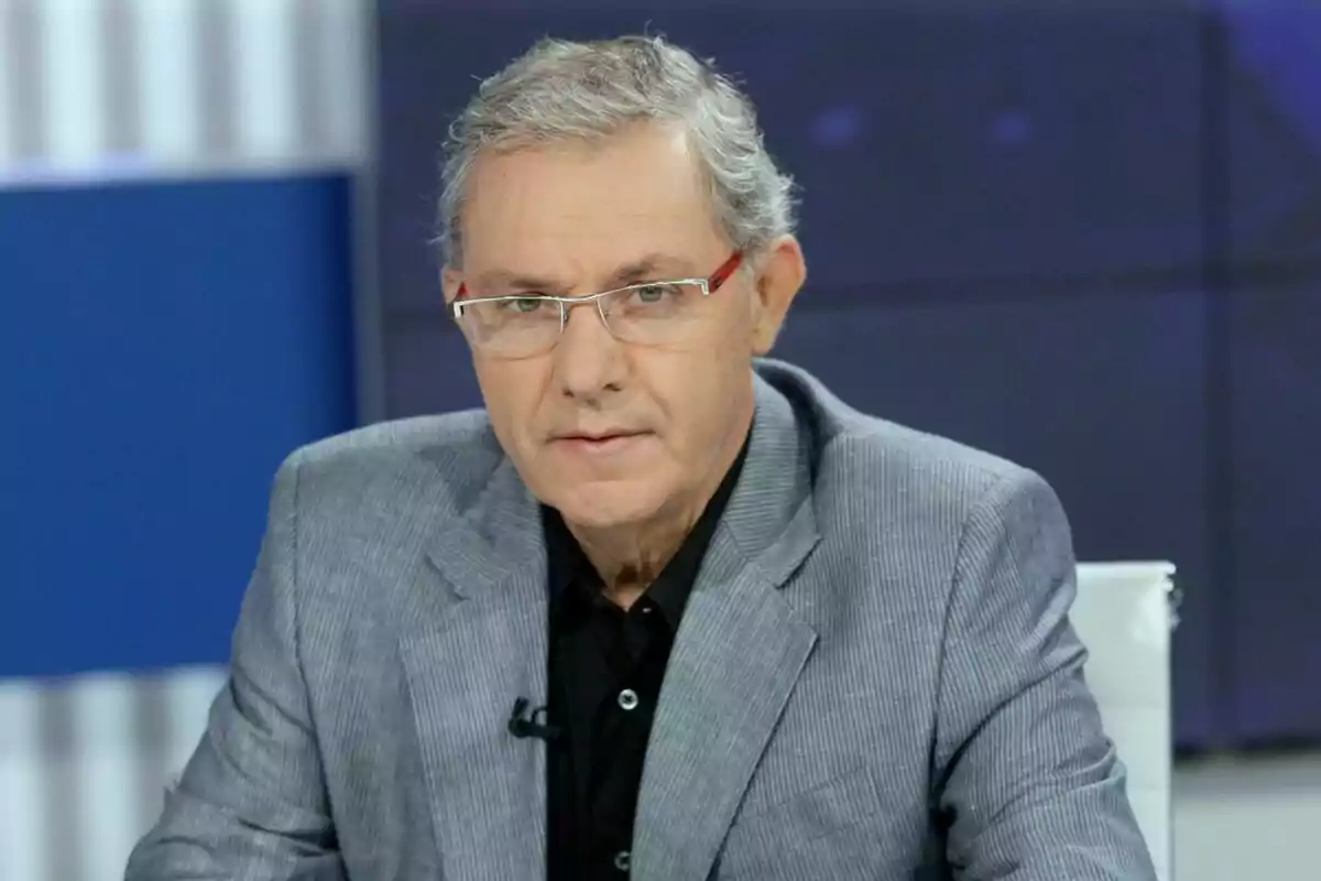 José Ángel de la Casa amb ulleres i cabell canós porta un vestit gris i camisa negra mentre està assegut en un estudi de televisió.